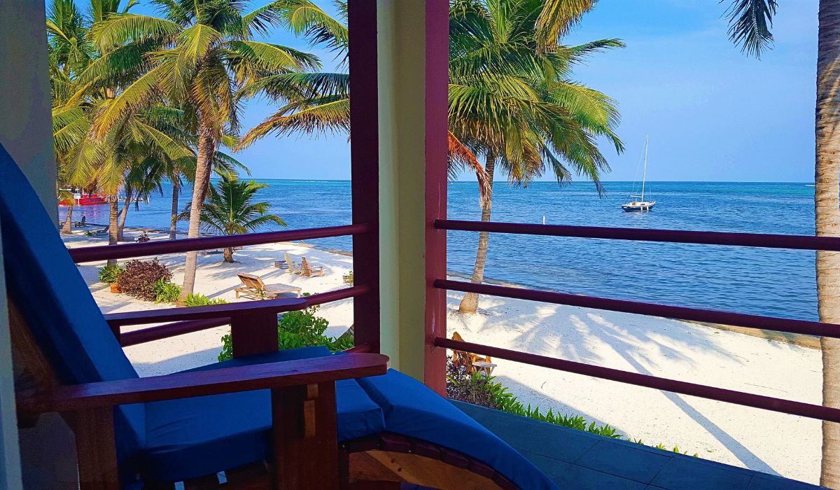 A tranquil view from a beachfront balcony in San Pedro, Belize, featuring a comfortable chair overlooking the sandy beach, swaying palm trees, and the calm Caribbean Sea with a sailboat anchored offshore.