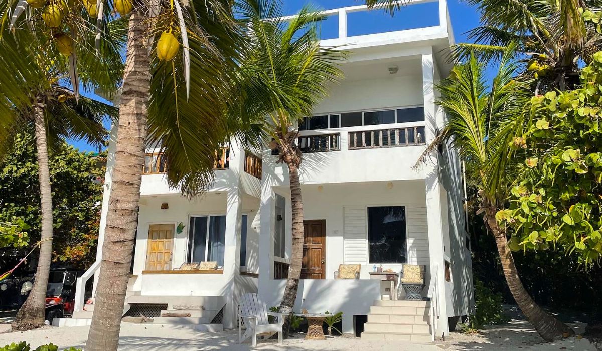 A charming beachfront villa in San Pedro, Belize, surrounded by palm trees and tropical foliage. The white exterior and cozy outdoor seating area offer a peaceful retreat, making it an ideal accommodation option for visitors looking to relax by the Caribbean Sea.