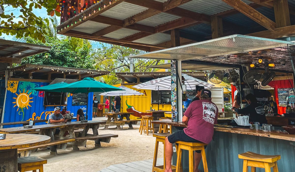 Visitors enjoy food and drinks at The Truck Stop in San Pedro, Belize, a popular outdoor food court made from repurposed shipping containers. The vibrant atmosphere, with picnic tables and colorful umbrellas, makes it a must-visit spot.