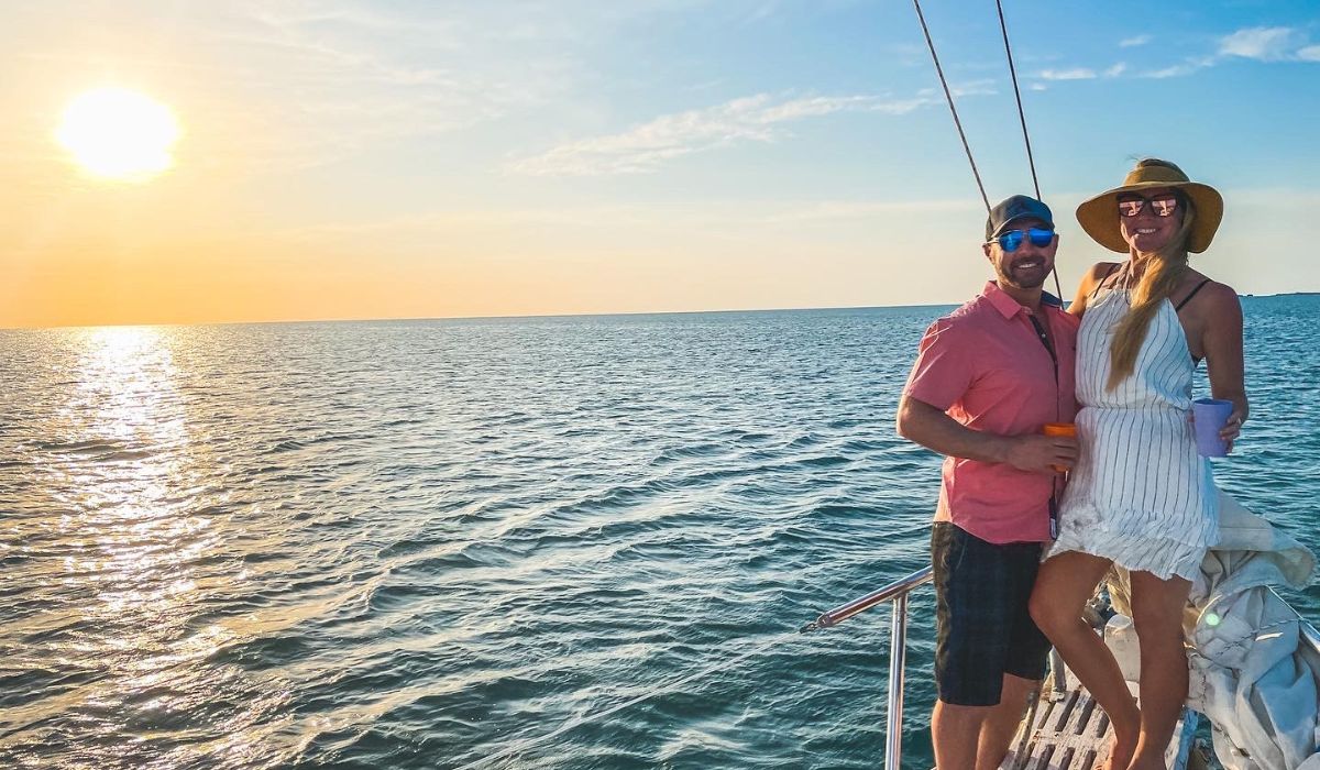 Kate and her husband enjoy a sunset tour on a boat off the coast of San Pedro, Belize. Kate is wearing a white dress and wide-brimmed hat, and her husband is dressed in a pink shirt and cap, smile as they stand at the bow of the boat, with the setting sun casting a golden glow over the water.