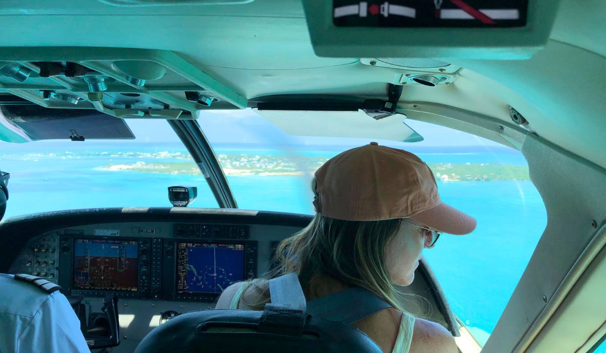 View from inside a small airplane cockpit as it flies over clear turquoise waters approaching San Pedro, Belize. Kate wearing a tan baseball cap is seated beside the pilot, looking out over the vibrant landscape below.