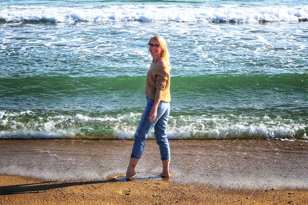 Kate walks barefoot along the Maine shoreline, with gentle waves rolling in behind her. She is wearing a light brown sweater and jeans, smiling as she enjoys the beach. The green-tinted water and sandy shore create a peaceful coastal scene, capturing a relaxing moment by the ocean.