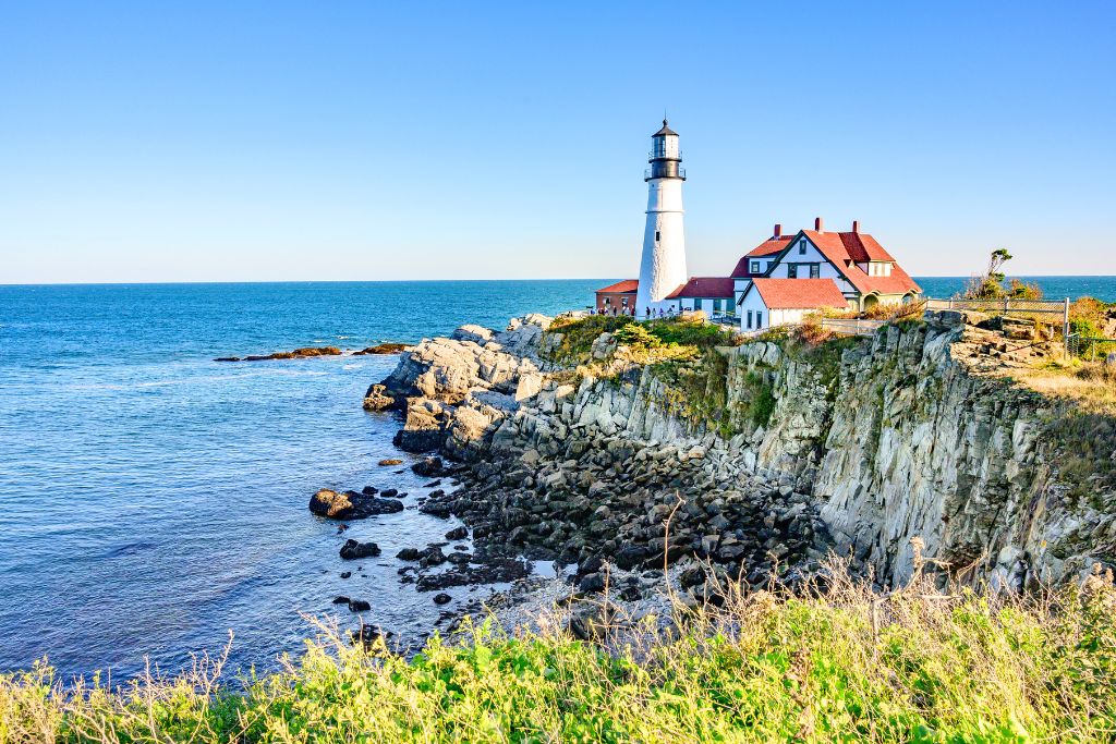 A scenic view of the Portland Lighthouse perched on a rock Maine coastline, with the blue waters of the Atlantic Ocean stretching into the horizon. The lighthouse has a white tower and red-roofed buildings, set against a clear blue sky. This is a picturesque coastal scene along a Boston to Bar Harbor Maine drive.