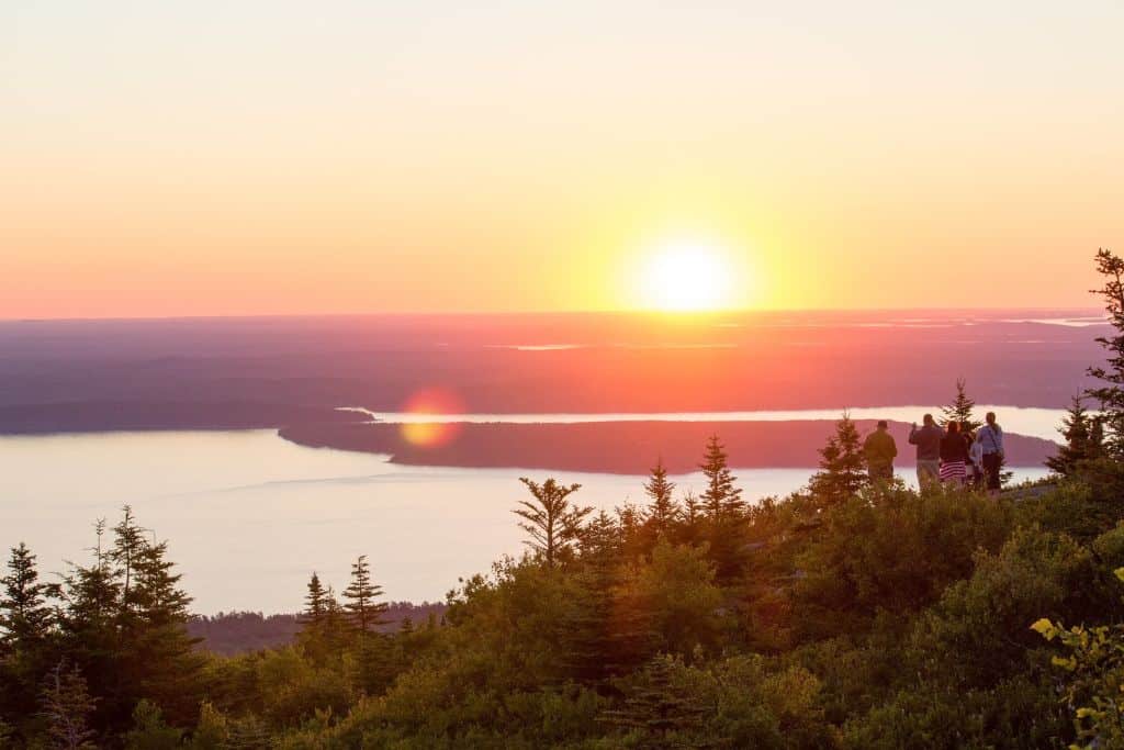 Photo of a hilltop at sunrise, admiring the view from Cadillac Mountain in Acadia National Park. The sun rises over the horizon, casting a warm glow over the surrounding trees and distant waters, creating a serene and breathtaking landscape.