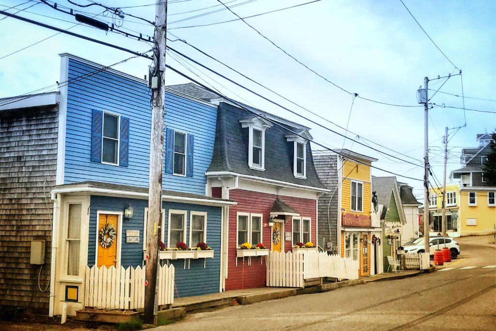 A row of colorful, charming houses lines a sloping street in Stonington, Maine, with buildings painted in shades of blue, red, and yellow. White picket fences and flower boxes adorn the storefronts, adding a cozy, welcoming feel. The scene captures the quaint character of a small New England town.