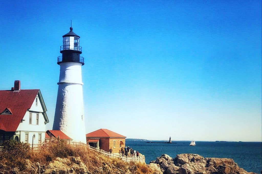 The iconic Portland Head Light stands tall on a rocky cliff overlooking the Atlantic Ocean in Portland, Maine. A bright blue sky and calm waters frame the scene, with a sailboat and another distant lighthouse visible on the horizon. The red-roofed lighthouse buildings add a classic coastal charm to this popular New England destination.