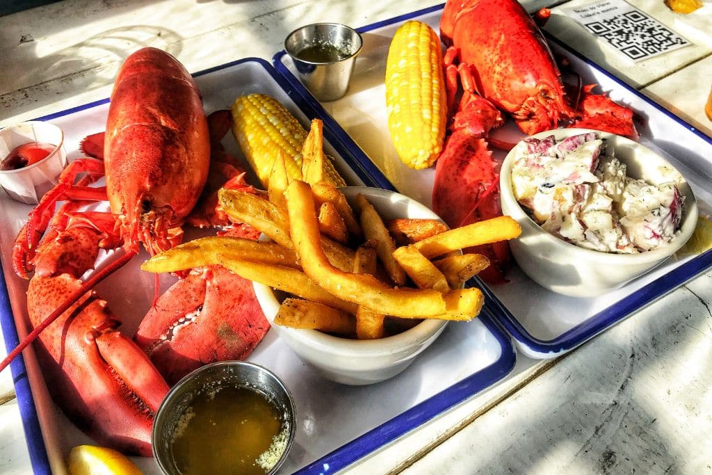 Two trays featuring traditional Maine lobster meals with whole boiled lobsters, corn on the cob, and sides of French fries and potato salad. A small dish of melted butter is included for dipping, along with a lemon wedge and a side of cocktail sauce. The bright, fresh seafood spread reflects a quintessential dining experience in coastal Maine.