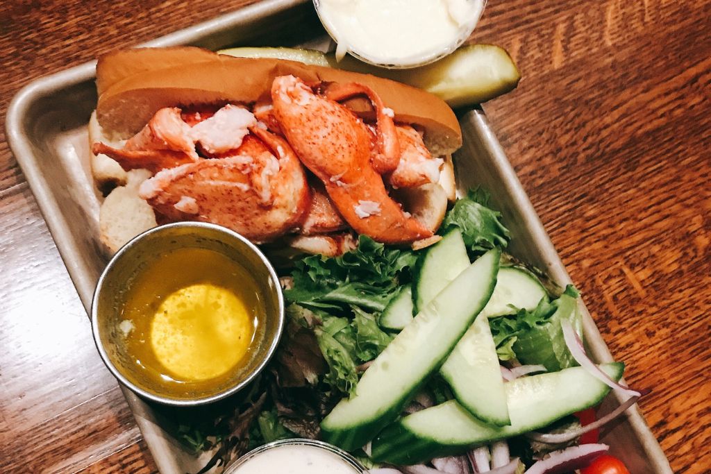 A lobster roll served on a toasted bun, filled with fresh lobster meat, accompanied by a side salad with cucumbers, red onions, and greens. A small cup of melted butter and a pickle spear are placed next to the sandwich, offering a classic Maine seafood dish. The rustic wooden table adds to the cozy, casual dining setting.