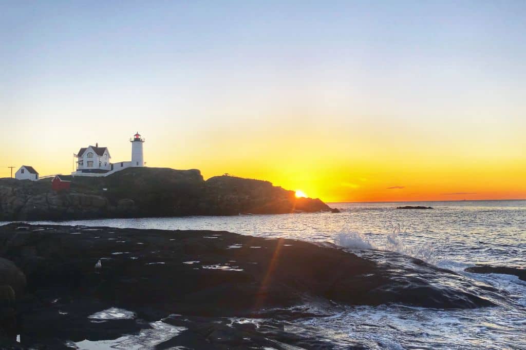 The sun sets behind the iconic Nubble Lighthouse, perched on a rocky outcrop overlooking the ocean. The sky glows with warm hues of orange and yellow, casting a soft light over the water and the rugged coastline. Gentle waves crash against the rocks in this peaceful coastal scene.