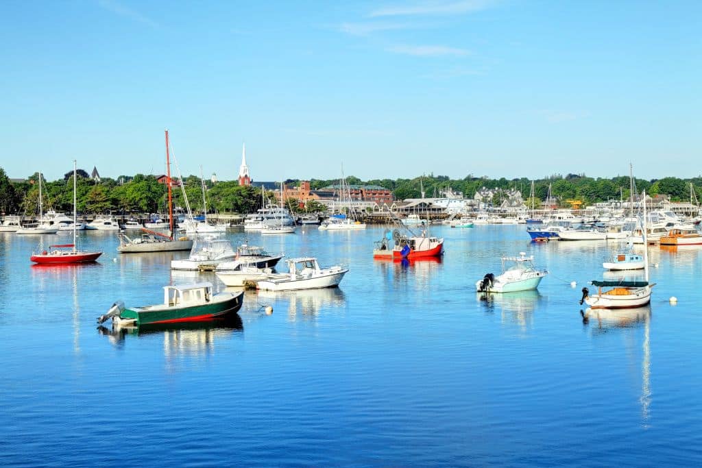 A peaceful harbor in Newburyport, MA, filled with small boats anchored on calm, blue waters under a clear sky. The shoreline features historic buildings, a church steeple, and lush greenery, reflecting the charm of this coastal New England town. The scene captures the serene beauty of a summer day by the water.