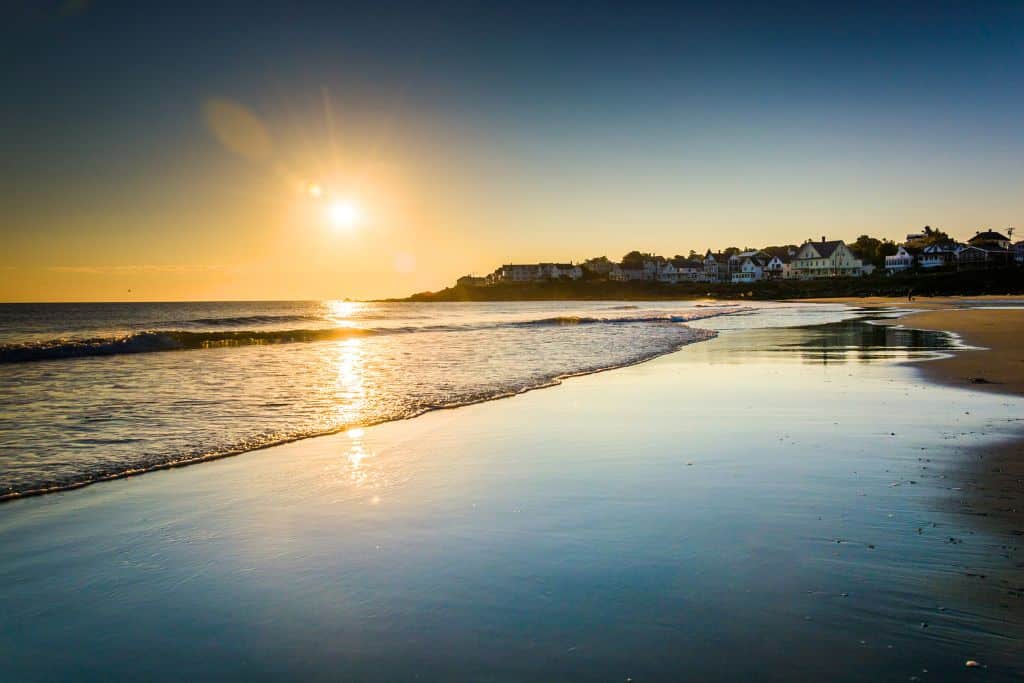 A serene sunset over a quiet beach in York,Maine, with the sun casting golden reflections on the wet sand and gentle waves lapping the shore. In the distance, charming seaside houses line the coast, basking in the warm evening light. The peaceful scene captures the beauty of a coastal New England town at dusk.