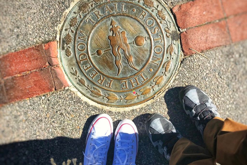 Two people stand beside a bronze plaque embedded in the sidewalk marking the Boston Freedom Trail. The plaque features an arrow and the trail's name, surrounded by decorative leaves. One person wears white sneakers, and the other wears black and gray shoes, adding a personal touch to this historic landmark in Boston.