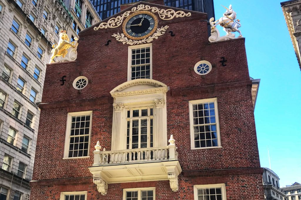 The historic Old State House in Boston, a red-brick building adorned with white trim and gold sculptures of a lion and a unicorn on its rooftop corners. A large clock is centered above the main balcony, with tall windows flanking both sides. The building stands among modern high-rise structures, blending colonial architecture with the surrounding cityscape.