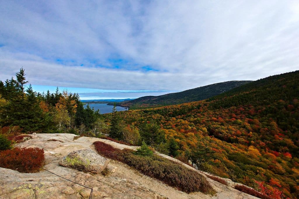 A stunning view from a rocky overlook in Acadia National Park, showcasing a vibrant autumn landscape. The rolling hills are covered in a mix of green, orange, and red foliage, leading down to a distant lake under a partly cloudy sky. The scenery highlights the park's natural beauty during the fall season.