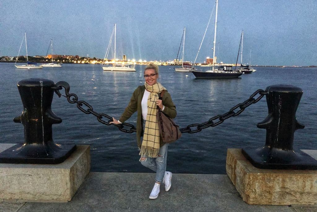 Kate poses by a large chain and bollards on the edge of the Boston waterfront, with sailboats anchored in the harbor behind her. She is dressed warmly in a scarf, jacket, and white sneakers as the lights from the city glow in the distance, reflecting off the calm evening waters. The scene captures a peaceful moment at dusk by the harbor.