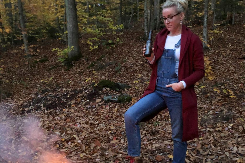 Kate, dressed in overalls, a red cardigan, and glasses, stands in a wooded area next to a camp fire during autumn, holding a drink. She rests one foot on a rock near a campfire, with fallen leaves covering the forest floor. The setting suggests a cozy outdoor camping evening in the fall, surrounded by nature.