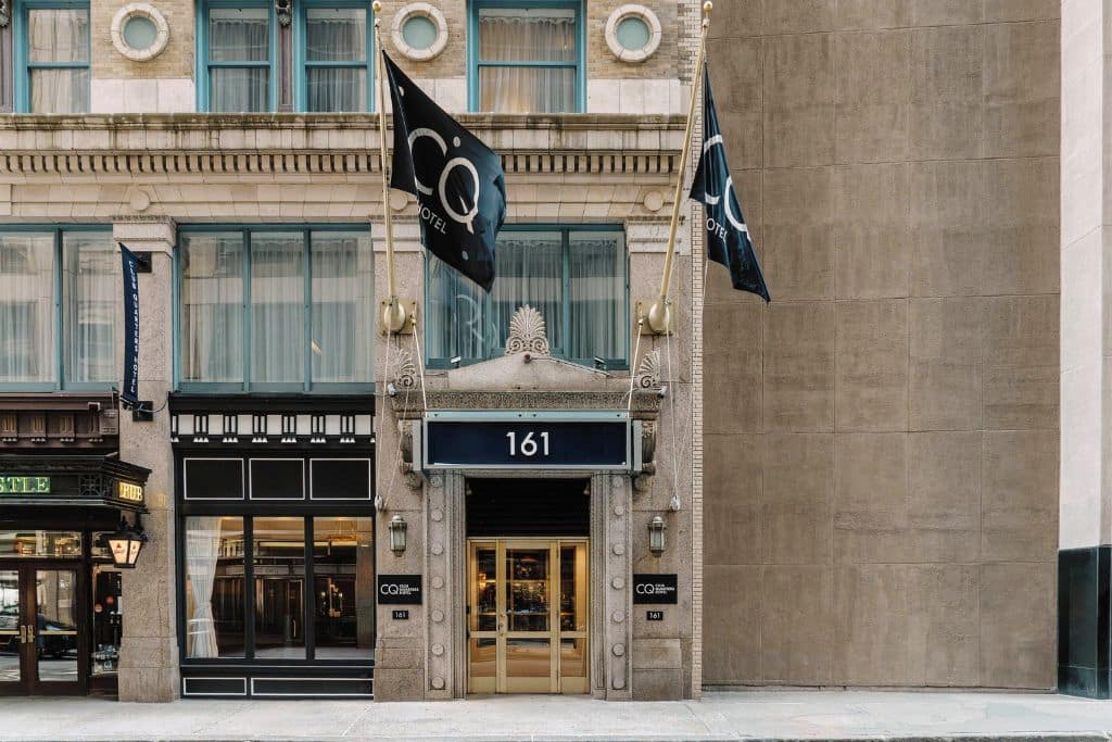 Entrance to the Club Quarters Hotel in downtown Boston, featuring a classic facade with large windows, flags bearing the hotel’s logo, and the address '161' prominently displayed above the doorway