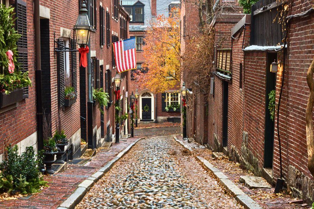 A cobblestone street lined with historic red-brick buildings, decorated with greenery. An American flag hangs above the street, and gas lamps line the sidewalk. The fall leaves scattered on the ground and a tree with autumn foliage add warmth to this picturesque alleyway, in Boston's Beacon Hill neighborhood.