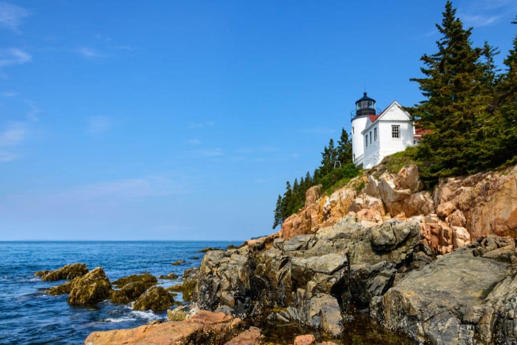 A picturesque white lighthouse stands atop a rocky cliff overlooking the blue waters of the Atlantic Ocean. Surrounded by evergreen trees, the lighthouse provides a striking contrast against the clear blue sky in Acadia National Park. The rugged coastline and calm sea complete this classic New England coastal scene.