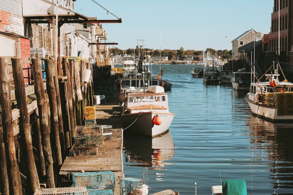 A narrow harbor filled with fishing boats docked alongside weathered wooden piers, with lobster traps stacked on the docks. The rustic waterfront buildings and calm waters reflect the busy, yet peaceful atmosphere of a working fishing village in Maine. The scene captures a typical day in a coastal town.