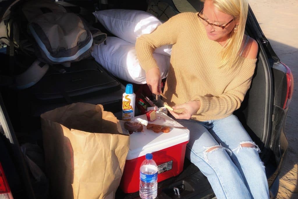 Kate sits at the back of an open car, preparing a snack with crackers and cheese. A red cooler, a paper bag, and a bottle of water are placed nearby, along with a can of Easy Cheese. The car's trunk is filled with travel essentials, including a backpack and pillows, suggesting a road trip or camping adventure.