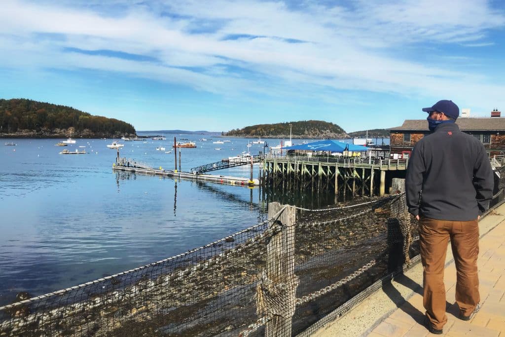 A man in a jacket and cap stands on a waterfront path in Bar Harbor, Maine, looking out over a calm bay dotted with small boats. Wooden docks and piers extend into the water, while forested islands and a blue sky with scattered clouds create a peaceful coastal scene. The setting reflects the charm of a quiet harbor town.