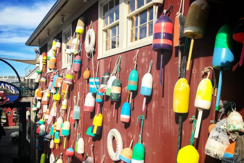 A red wooden building in Bar Harbor, Maine, is adorned with a colorful assortment of fishing buoys hanging on its exterior. The bright buoys in various shapes and patterns add a vibrant and coastal charm to the rustic setting, with a clear blue sky visible in the background. The scene captures the essence of a working fishing town.