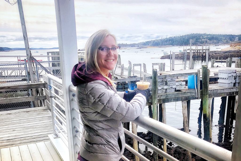 Kate in a light jacket enjoys a warm drink while standing on a dock overlooking the harbor in Stonington, Maine. Fishing crates and wooden piers line the water, with small boats anchored in the distance against a backdrop of forested islands. The overcast sky adds to the quiet, coastal atmosphere of this scenic harbor town.