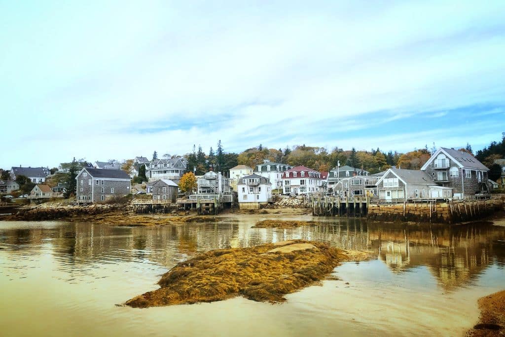 A view of charming seaside homes and buildings on stilts along the waterfront in Stonington, Maine. The houses, with their weathered gray exteriors, sit along the rocky shoreline with seaweed-covered rocks exposed during low tide. The scene captures the peaceful, rustic beauty of this coastal town under a partly cloudy sky.