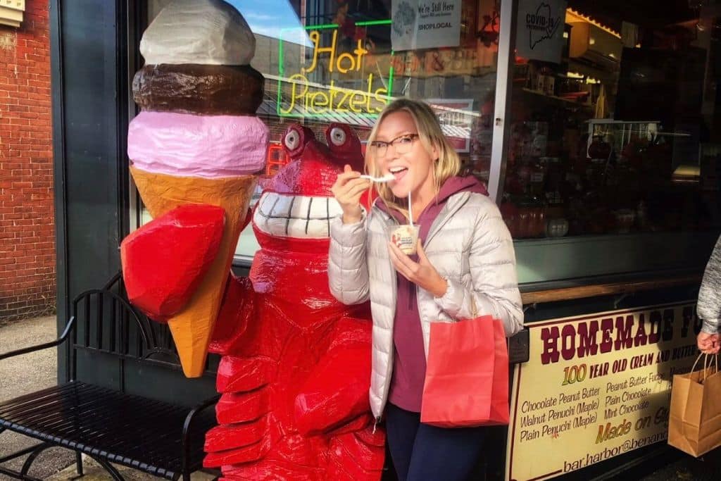 Kate in a light gray jacket, holding a small cup of ice cream and a red shopping bag, poses next to a bright red lobster statue holding an oversized ice cream cone outside a shop in Bar Harbor, Maine. The shop window features signs like "Hot Pretzels" and "Homemade Fudge."