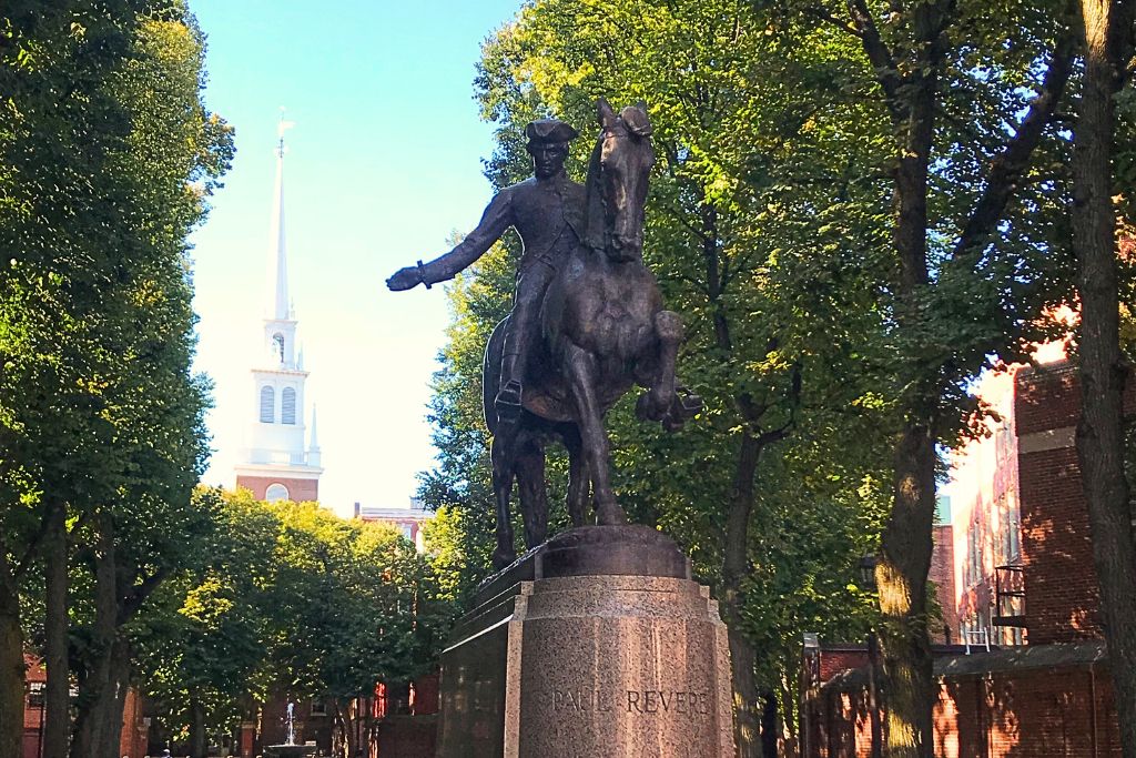 Statue of Paul Revere on horseback in Boston's historic North End, set among tall trees with the white steeple of the Old North Church visible in the background, commemorating his famous midnight ride during the American Revolution