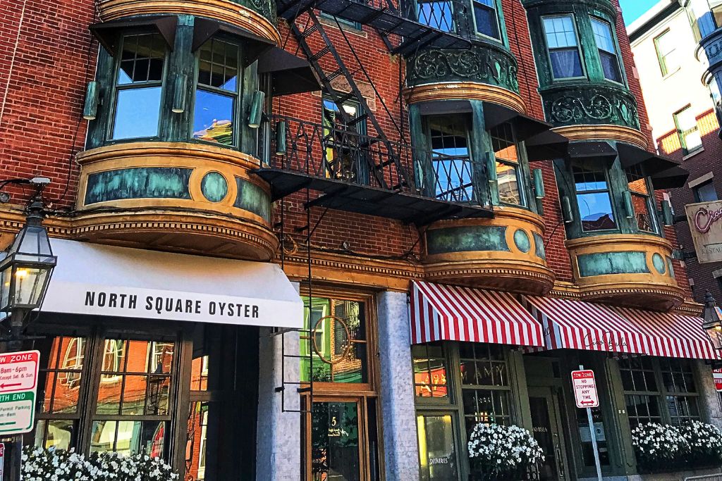 Exterior of North Square Oyster in Boston's North End, featuring a historic red-brick building with large, ornate bay windows framed in green copper and striped awnings over the entrance, creating a classic and inviting look.