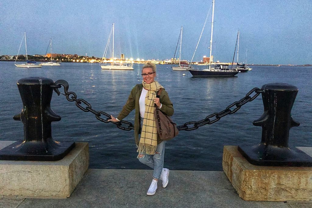 Kate standing by the Boston Harbor at dusk, holding onto a large chain connected to mooring posts, with sailboats floating on the calm water and the city skyline illuminated in the background.