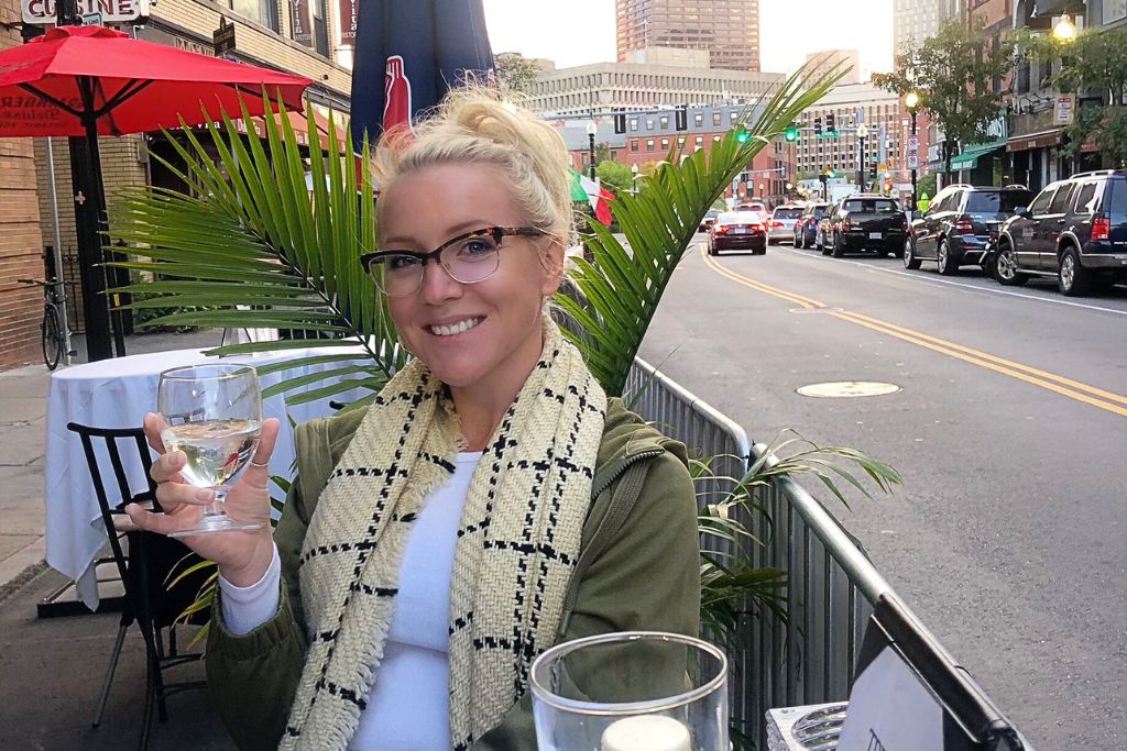 Kate sitting at an outdoor restaurant in Boston, smiling and holding a glass of white wine, with city streets, cars, and buildings in the background as the sun sets.