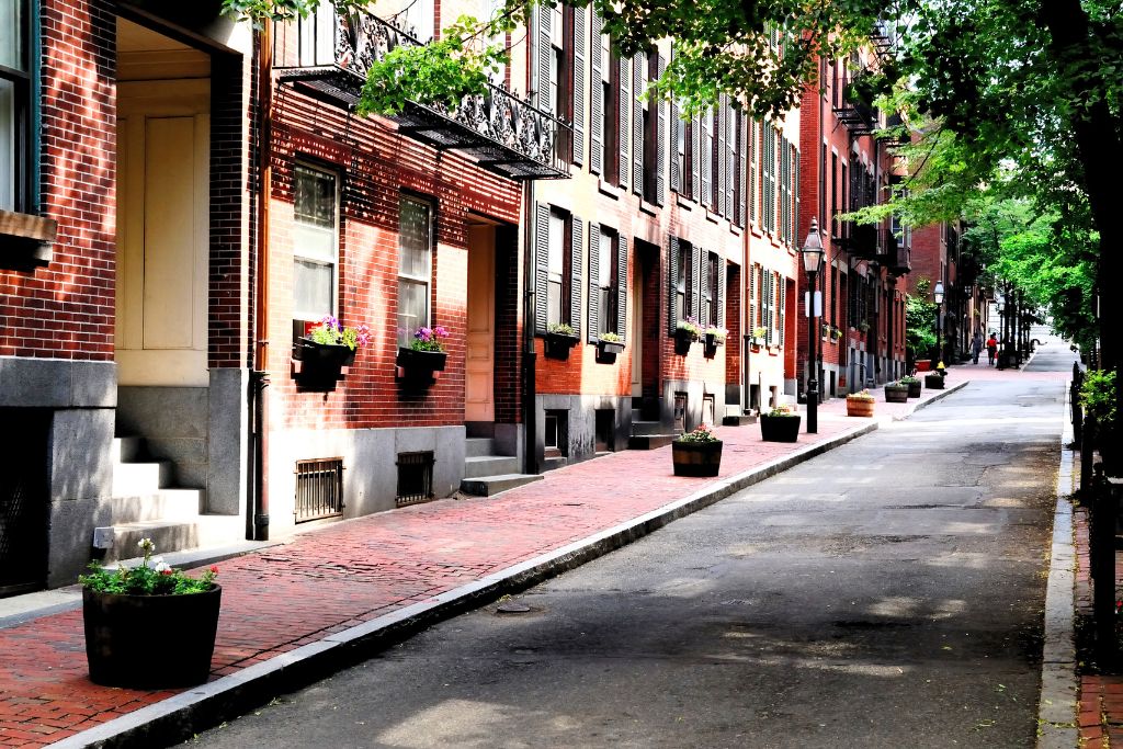 Charming, narrow street in Boston lined with historic red-brick townhouses, decorated with flower boxes and shaded by green trees, showcasing the city's picturesque residential architecture and peaceful urban setting.