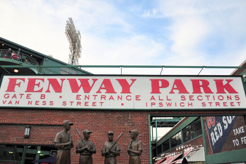 Entrance to Boston's iconic Fenway Park, featuring a large sign with red lettering above statues of legendary baseball players. The historic ballpark is a must-visit destination for Red Sox fans and sports enthusiasts.