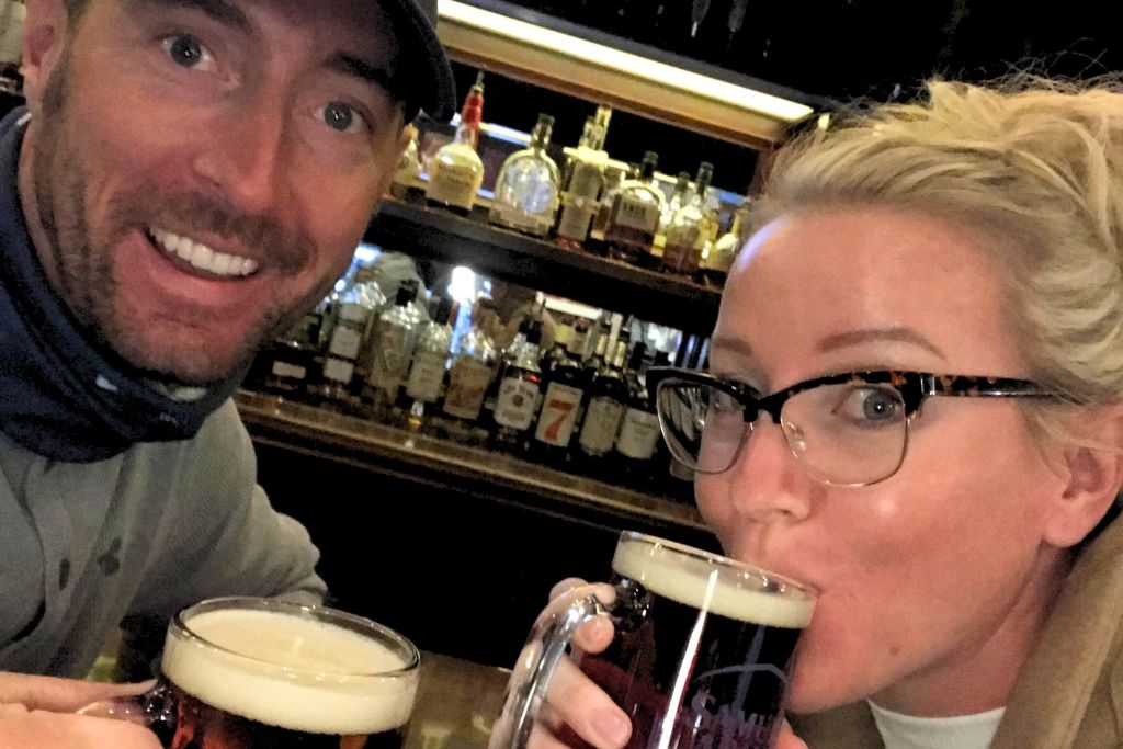 Kate and her husband enjoying a pub crawl in Boston, smiling as they hold mugs of beer in front of a bar lined with various bottles of liquor, capturing a fun moment during their city adventure.