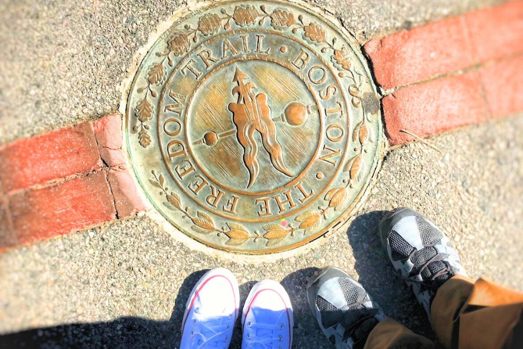 Bronze medallion embedded in the sidewalk marking the Freedom Trail in Boston, with two people’s feet, one in white sneakers and the other in black, standing beside it. The medallion signifies the historic walking trail through Boston's important landmarks.