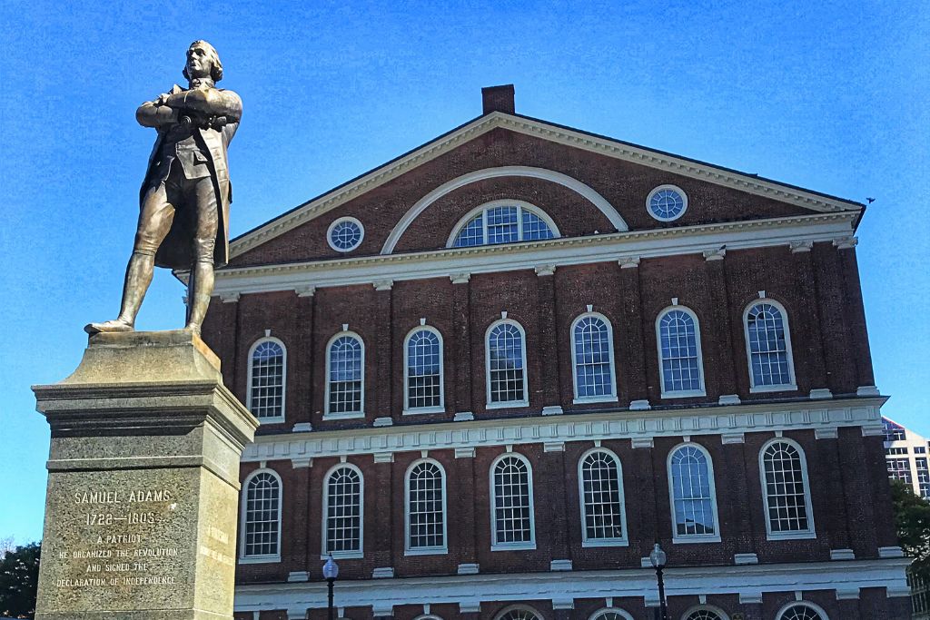 Statue of Samuel Adams in front of Faneuil Hall in Boston, with a clear blue sky in the background. The statue, standing on a tall pedestal, honors Adams as a Patriot who organized the revolution and signed the Declaration of Independence.