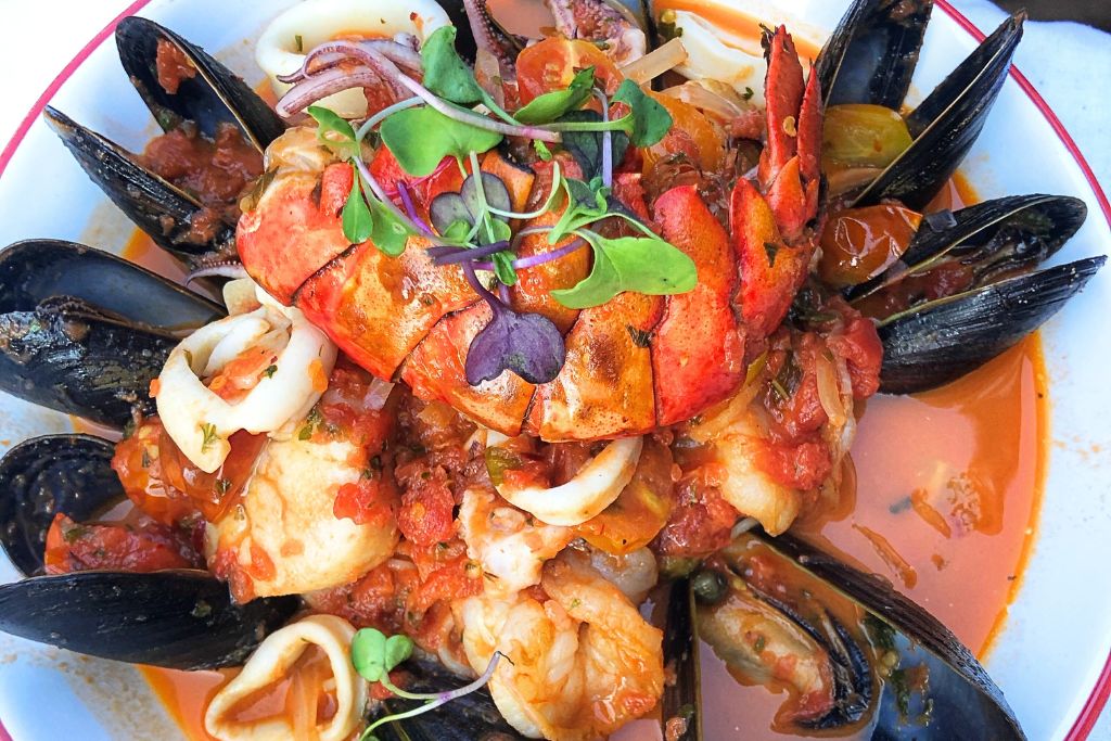 Close-up of a seafood dish featuring lobster, mussels, squid, and other shellfish in a rich tomato-based broth, garnished with microgreens. A delicious representation of Boston's famous seafood cuisine.