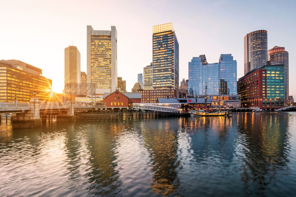 Scenic view of the Boston skyline at sunset with modern glass skyscrapers and historic waterfront buildings reflecting in the harbor, showcasing Boston's blend of history and modern architecture. Is Boston worth visiting for its iconic cityscape and waterfront attractions? Absolutely.