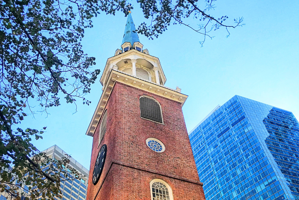 Upward view of the Old South Meeting House in Boston, featuring its tall brick tower and clock, framed by tree branches and modern glass buildings in the background, symbolizing the city's blend of historical and contemporary architecture.