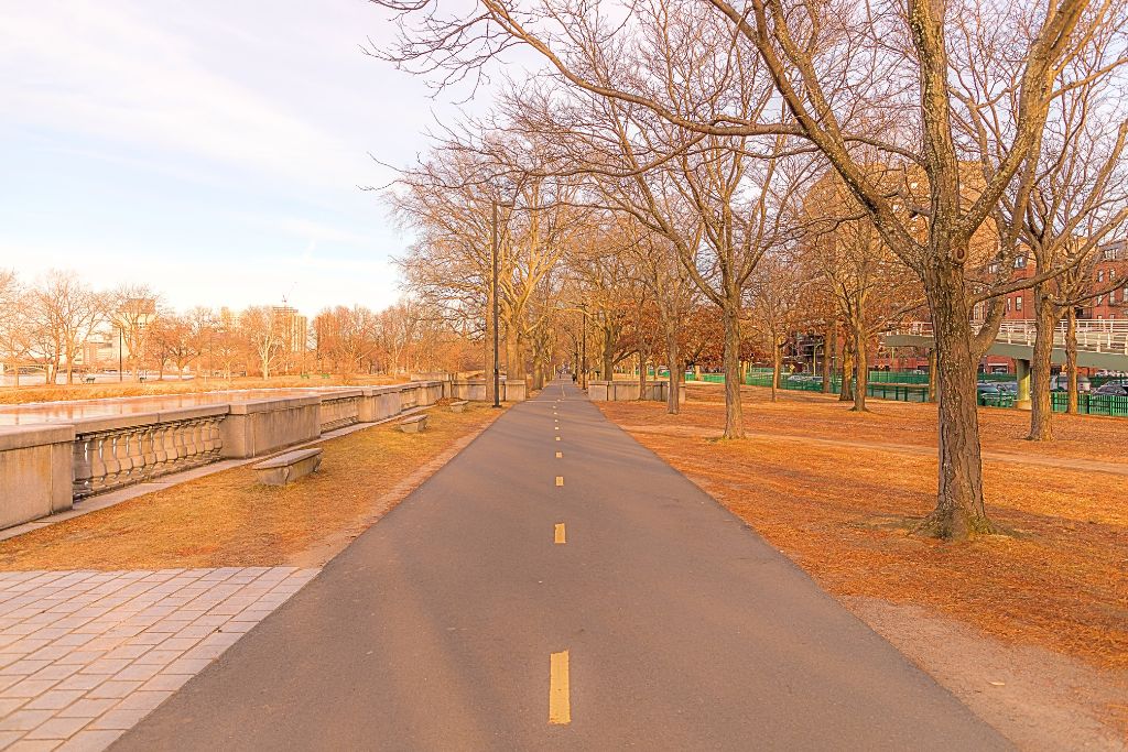 Empty pathway along the Charles River Esplanade in Boston during late autumn, with bare trees lining the trail. The warm light of the setting sun casts a golden hue over the scene, creating a peaceful atmosphere.