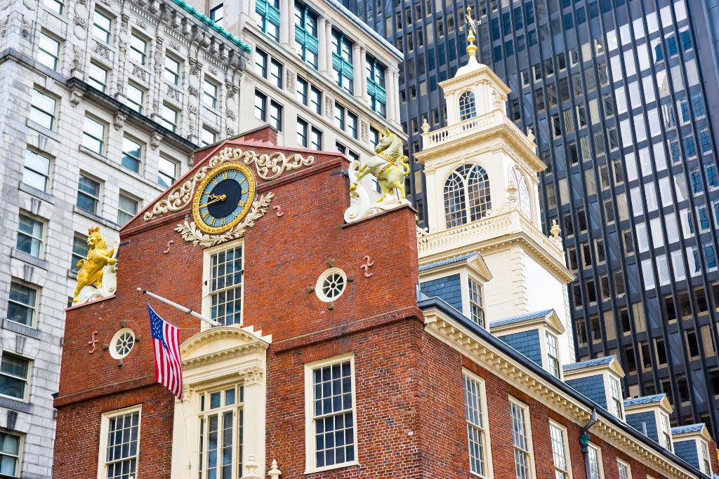 Close-up view of the Old State House in Boston, a historic red brick building adorned with a large clock and golden statues of a lion and unicorn. An American flag is displayed near the entrance, and modern skyscrapers surround the colonial-era structure.