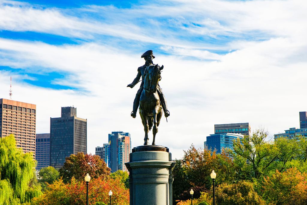 Statue of George Washington on horseback in the Boston Public Garden, with the city skyline in the background. The park's fall foliage contrasts with the modern buildings under a partly cloudy sky.