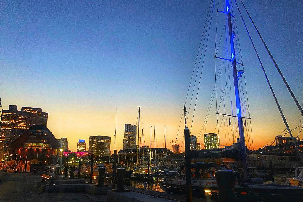 Boston Harbor at sunset, featuring sailboats docked in the marina with the city skyline illuminated in the background. The sky transitions from warm orange near the horizon to deep blue above, creating a peaceful waterfront evening scene.