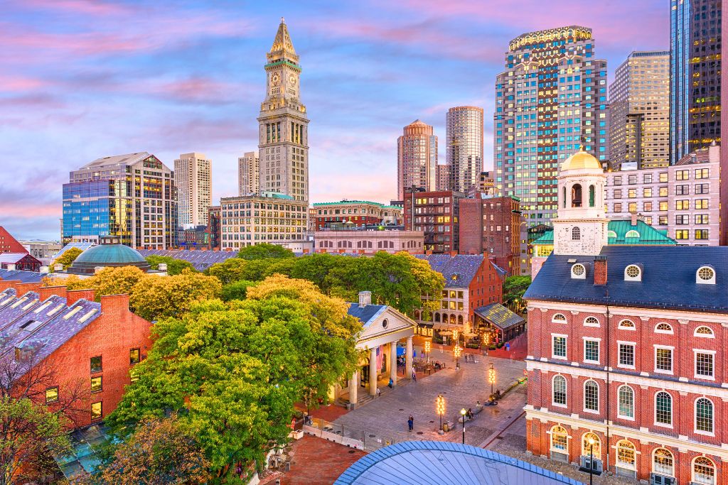 This image shows the iconic Faneuil Hall and Quincy Market in Boston, with the Custom House Tower and modern skyscrapers in the background. The photo captures a blend of historic and contemporary architecture under a pastel evening sky, representing a vibrant part of downtown Boston, a must-visit area when exploring the city in October.