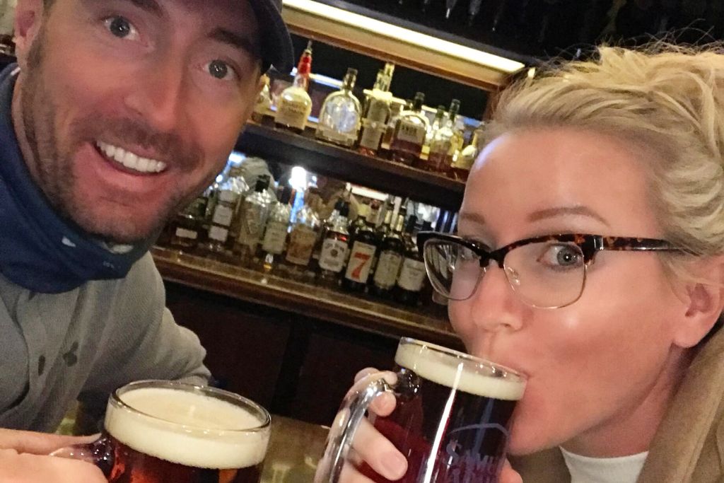 Kate and her husband enjoying beers at a bar during a pub crawl in Boston. Both are smiling, with Kate sipping from her glass while sitting in front of a shelf filled with bottles of liquor.