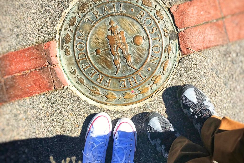 Bronze marker for Boston's Freedom Trail embedded in the sidewalk, with two pairs of feet visible, one in white sneakers and the other in gray, standing on either side of the brick-lined path. The marker reads 'The Freedom Trail Boston,' a popular historical site to explore, especially in October.