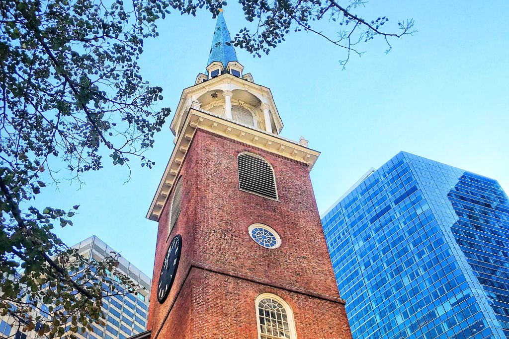 Upward view of the Old South Meeting House in Boston, featuring its historic red brick tower and blue steeple, framed by branches from nearby trees. The church stands in contrast to the surrounding modern glass skyscrapers, representing the city's blend of old and new architecture.